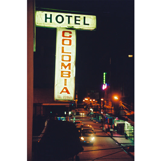 A vintage style photograph featuring a neon sign for 'HOTEL COLUMBIA' with a dark background, showing the hotel's facade and the surrounding area at night.