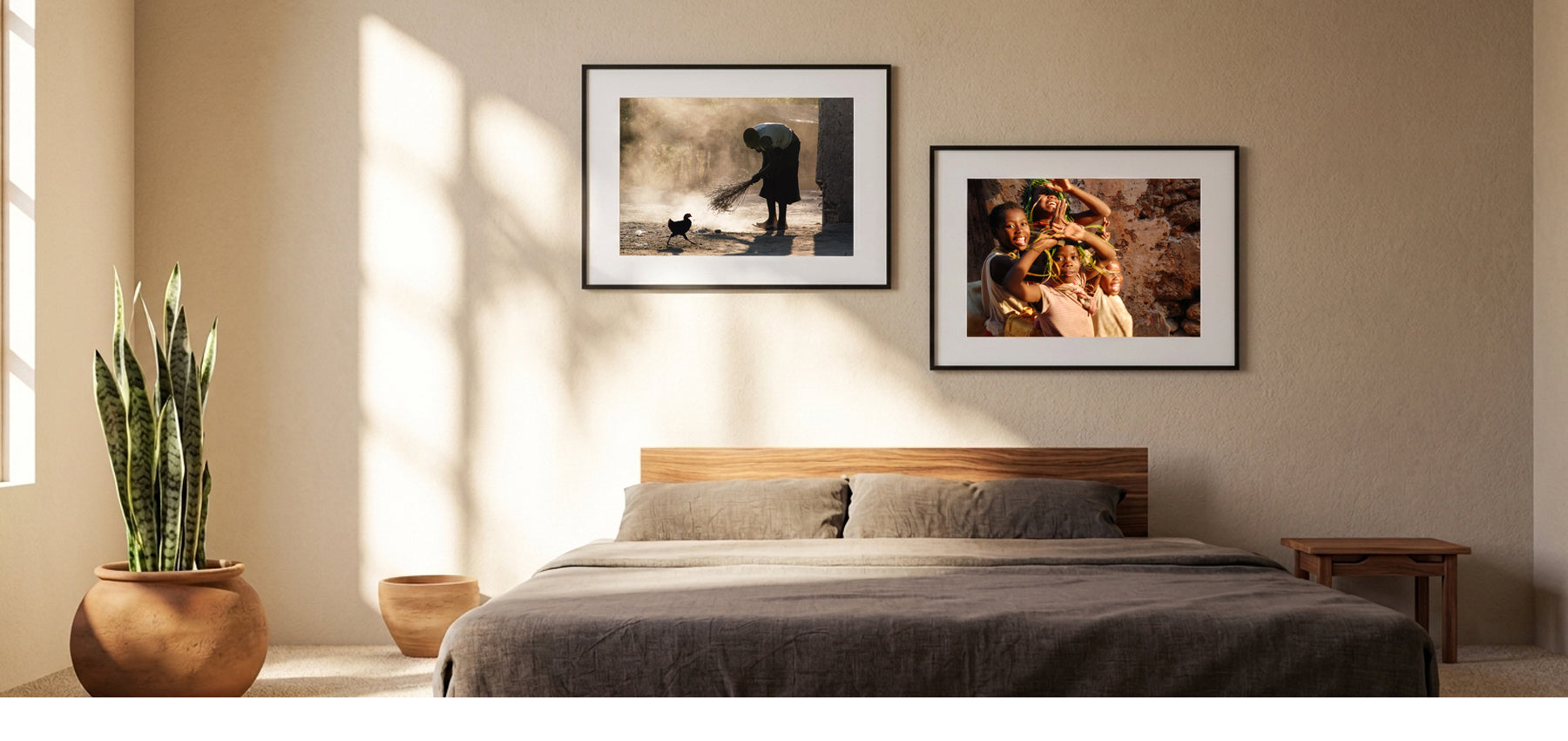 A styled bedroom interior with a bed, wall art, and a potted snake plant. Two framed photographic prints hang above the bed on a beige wall, with warm sunlight casting shadows across the room.