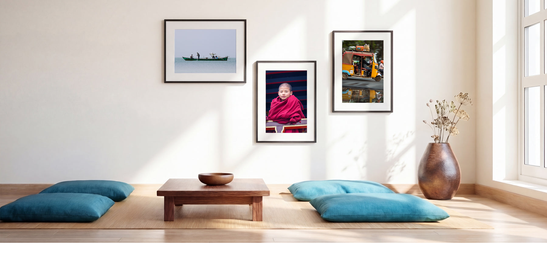 A styled interior room with three framed wall art prints hanging above a low wooden table and teal floor cushions. The prints depict a boat scene, a person in red robes, and a colorful street scene.