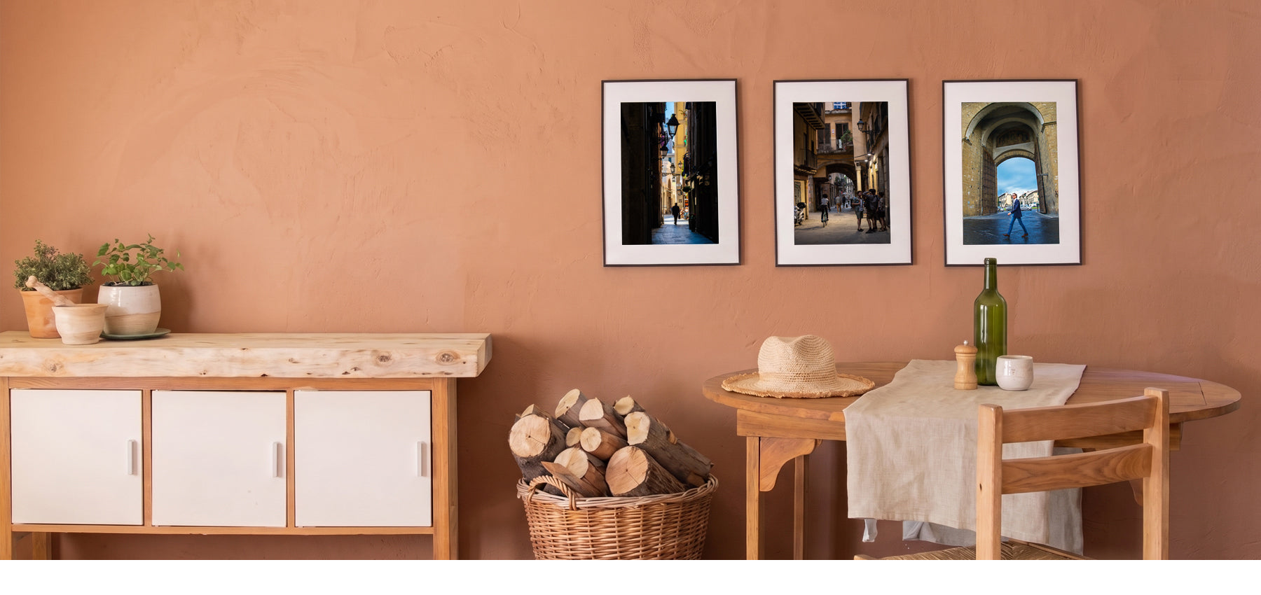 Three framed vertical art prints depicting scenic architectural street scenes, displayed on a terracotta-colored wall above a wooden sideboard and dining table.