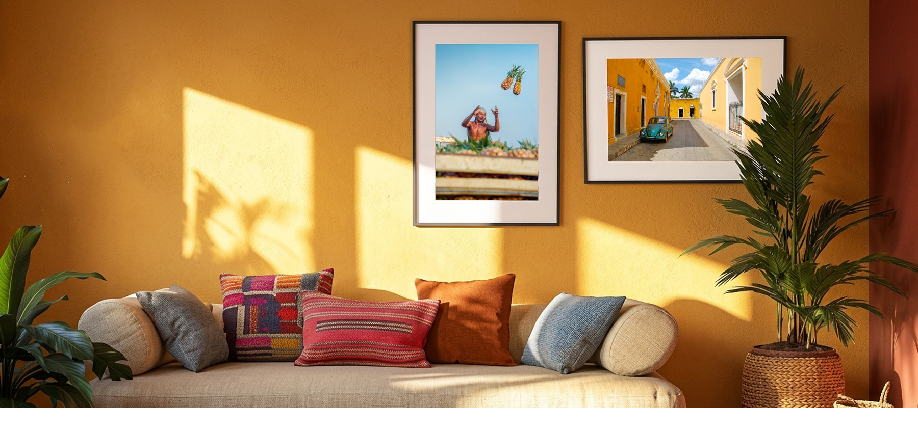 A styled interior scene with a beige sofa, colorful throw pillows, a potted plant, and two framed wall art prints hanging on a warm mustard-yellow wall. The prints depict colorful outdoor scenes, and the room is lit by strong diagonal sunlight with soft shadows.