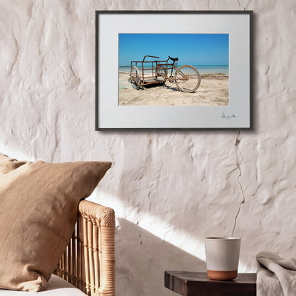 Framed photograph of an old cart on a beach wall-mounted above a textured wall with pillows and a cup.